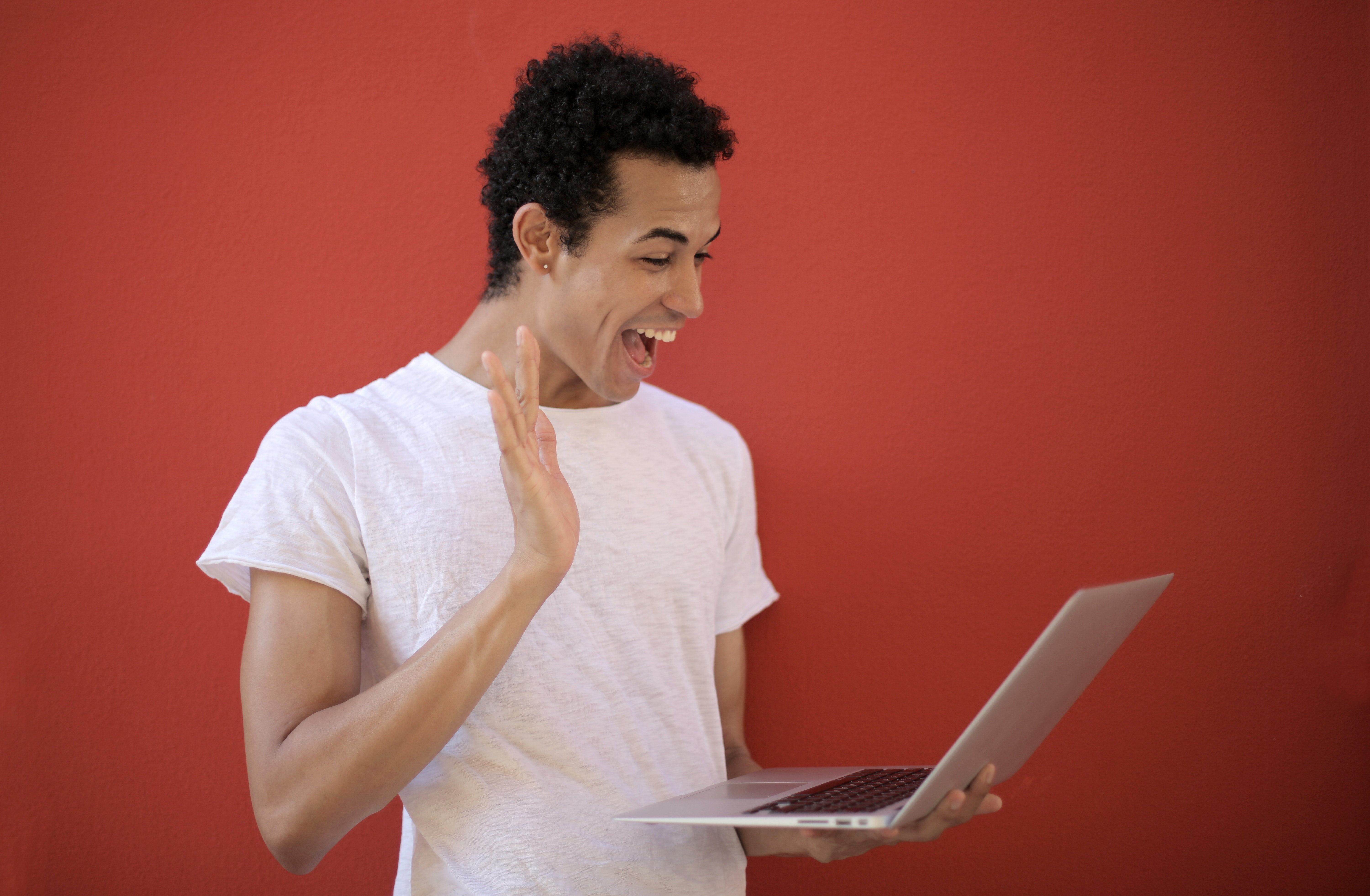 a guy waves happily at his laptop that he is holding in his other hand